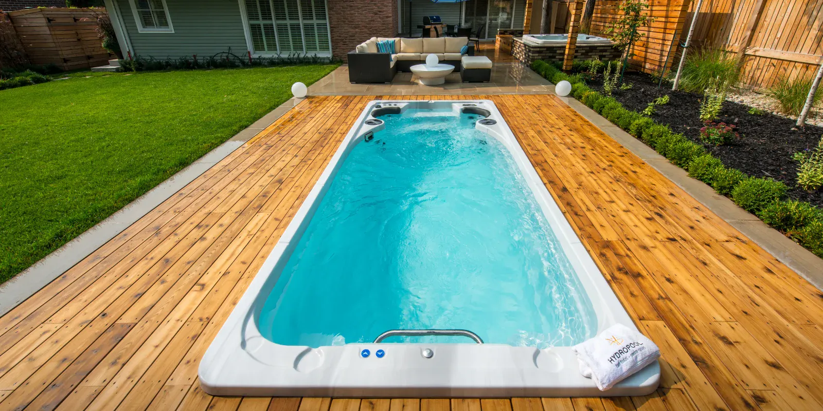 Hydropool swimspa nedsänkt i ett trädäck av ceder i en villaträdgård, med loungemöbler och spabad i bakgrunden