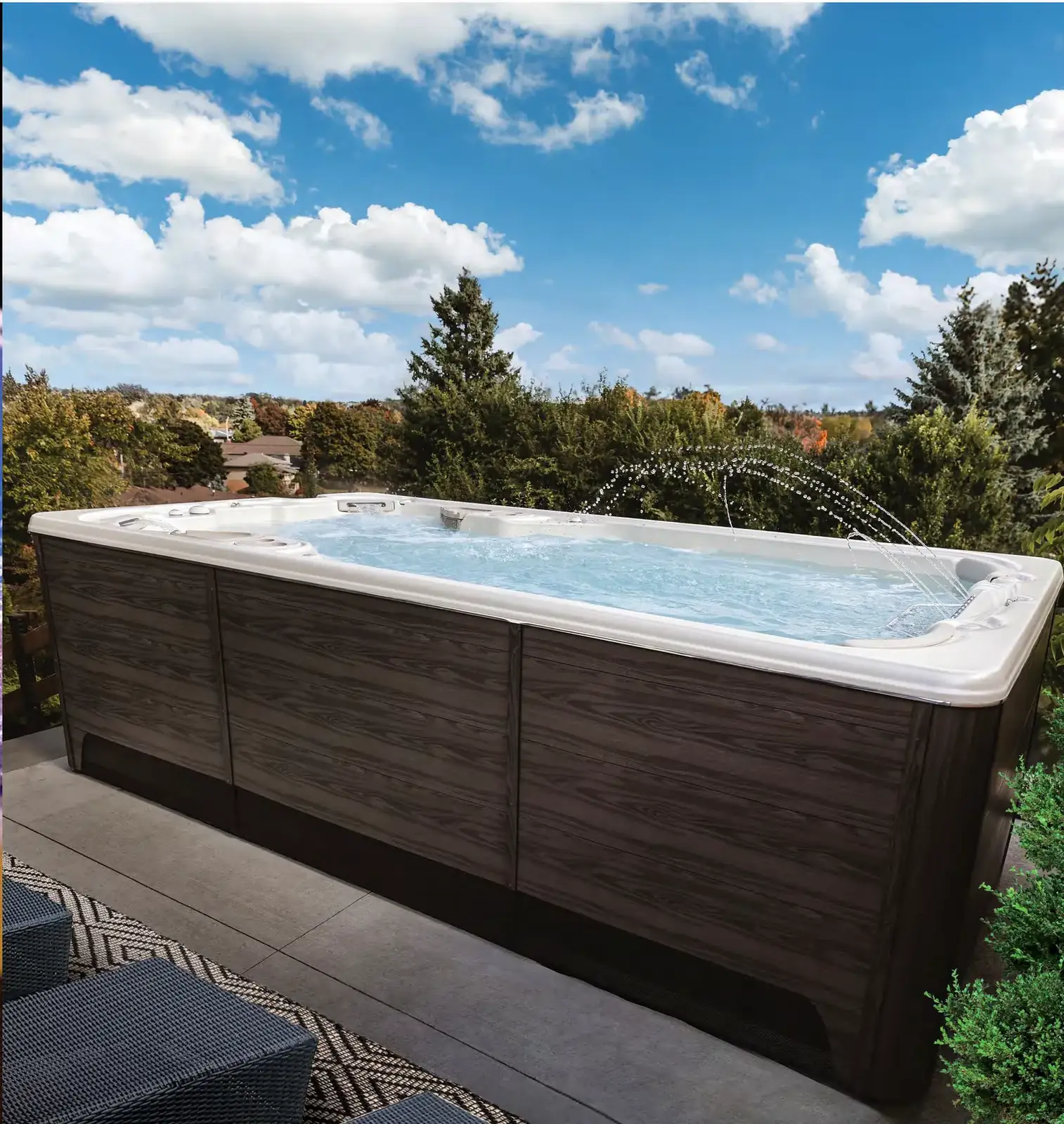 Hydropool swimspa med mörk träpanel på en terrass, med utsikt över trädtoppar och blå himmel